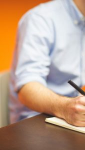Man at a desk writing in a notepad whilst using a computer.